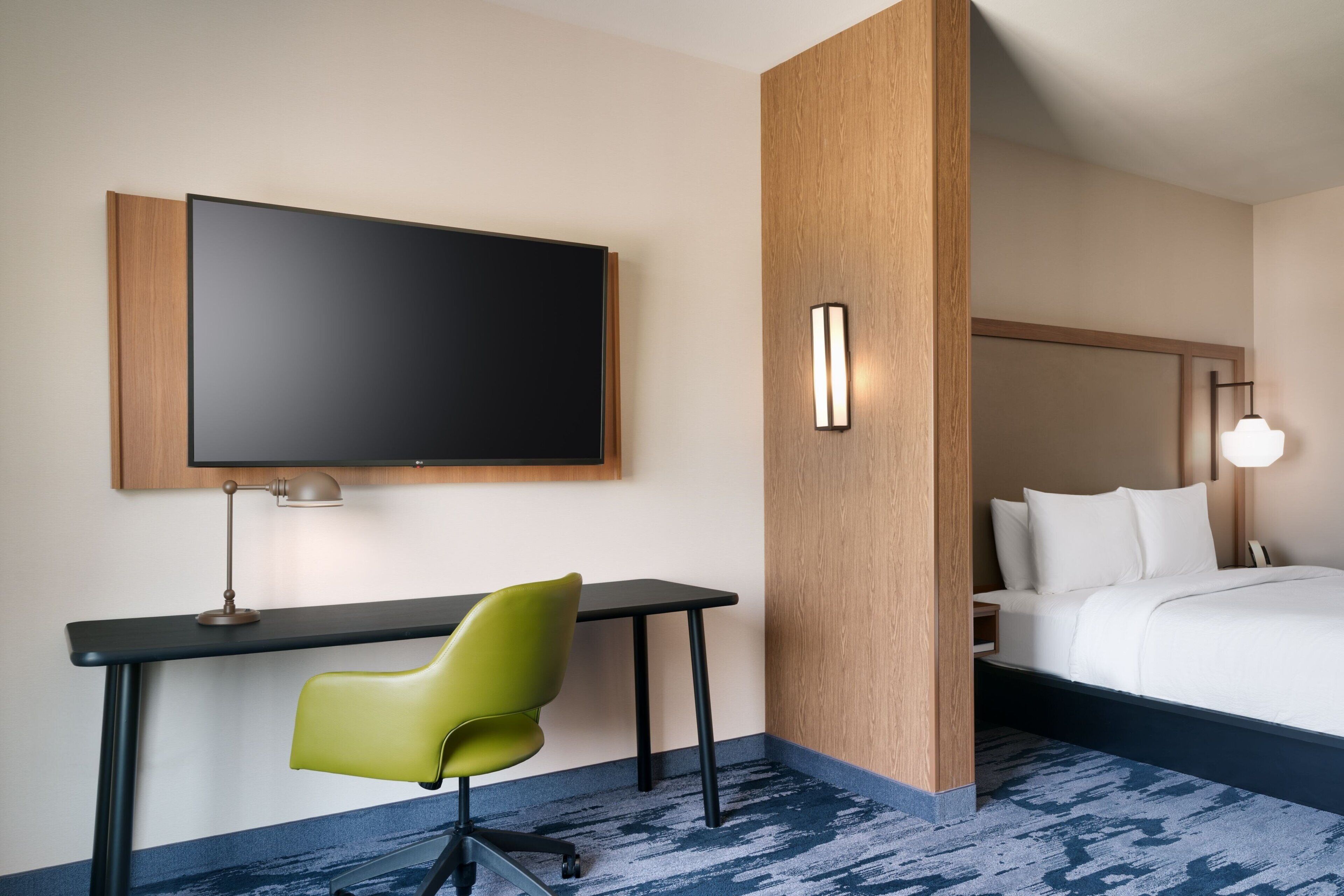 A hotel room with a bed, a desk with a chair, a flat-screen TV, and a wall-mounted light fixture.
