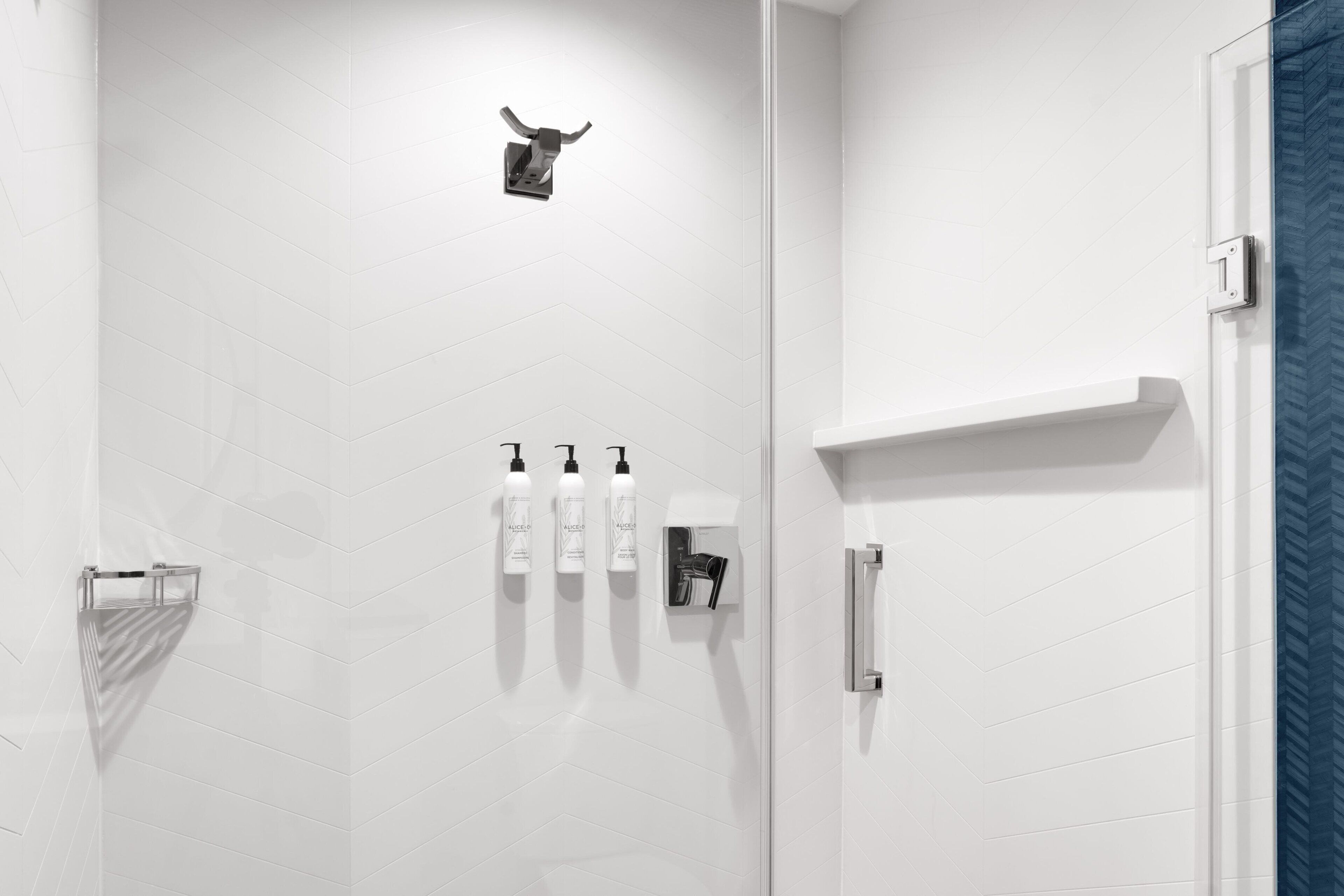 Un baño moderno con paredes de azulejos blancos, una ducha con puerta de vidrio y tres botellas de champú en un dispensador de pared.