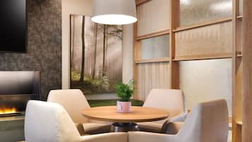 A modern living room with a round wooden table, beige armchairs, a flat-screen TV, a fireplace, and a large window with a forest view.