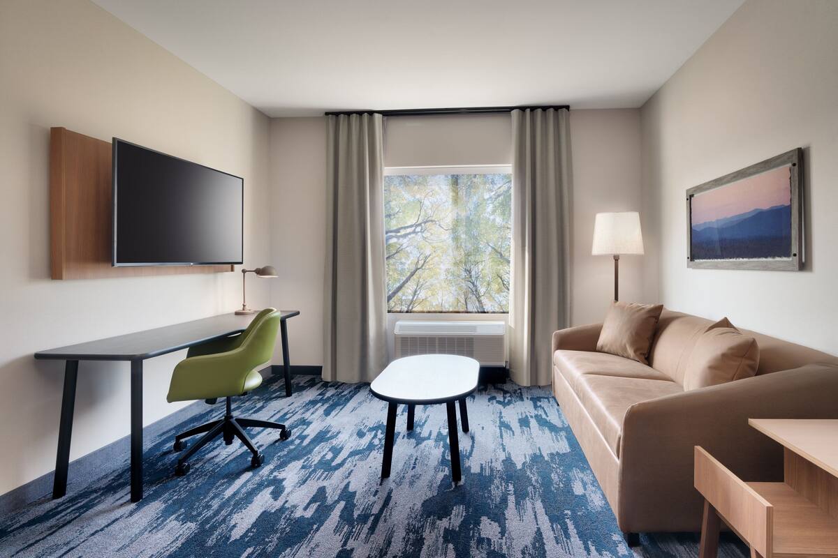 Une chambre d’hôtel équipée d’un téléviseur à écran plat, d’un bureau, d’une chaise verte, d’une table blanche, d’un canapé beige et d’une fenêtre donnant sur des arbres.