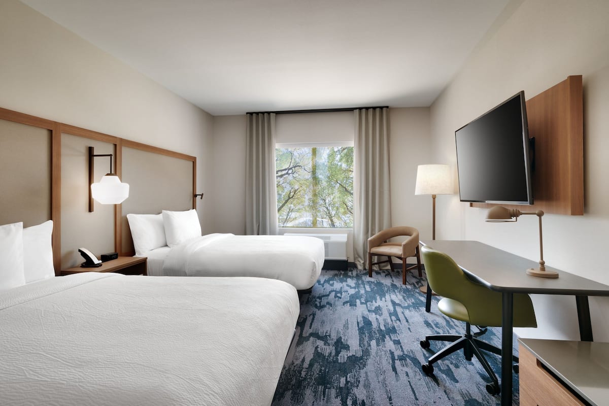 Une chambre d’hôtel avec deux lits, un bureau, une chaise, une télévision et une fenêtre donnant sur des arbres.