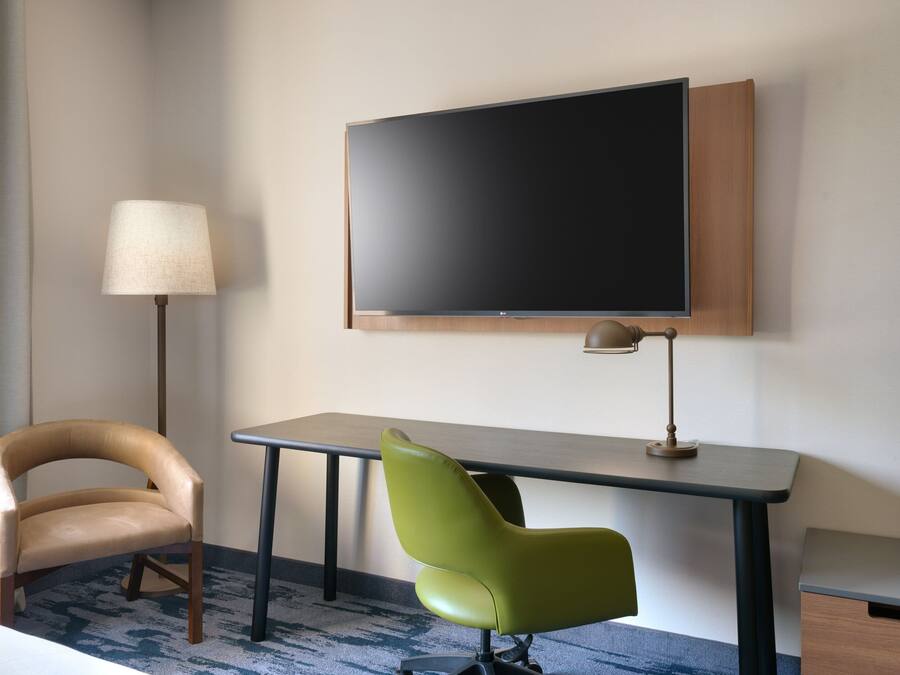 Ett hotellrum med en stor, platt-TV som är monterad på väggen, ett skrivbord med en lampa och två stolar, en beige och en grön.