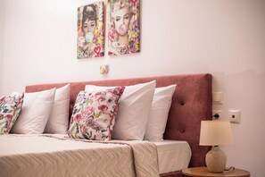 A neatly made bed with floral pillows, a round table with a lamp, and two wall art pieces.
