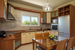 A modern kitchen with wooden cabinets, a stainless steel range hood, a built-in microwave, a refrigerator, a glass dining table, and a window with a view of greenery.