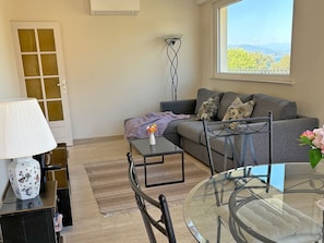 Un salon avec un canapé, une table basse en verre, une lampe et une vue sur la verdure par une fenêtre.