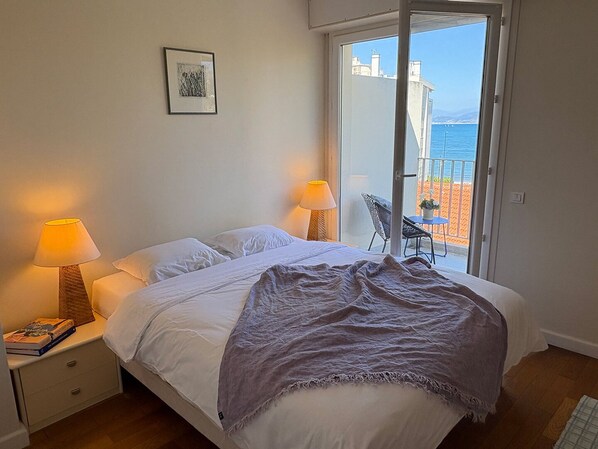 Une chambre à coucher avec un lit, deux lampes de chevet, une table de nuit, un tableau encadré au mur et une vue sur la mer par une porte-fenêtre coulissante.