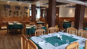 Un restaurante con mesas y sillas de madera, manteles verdes y plantas en macetas sobre las paredes.