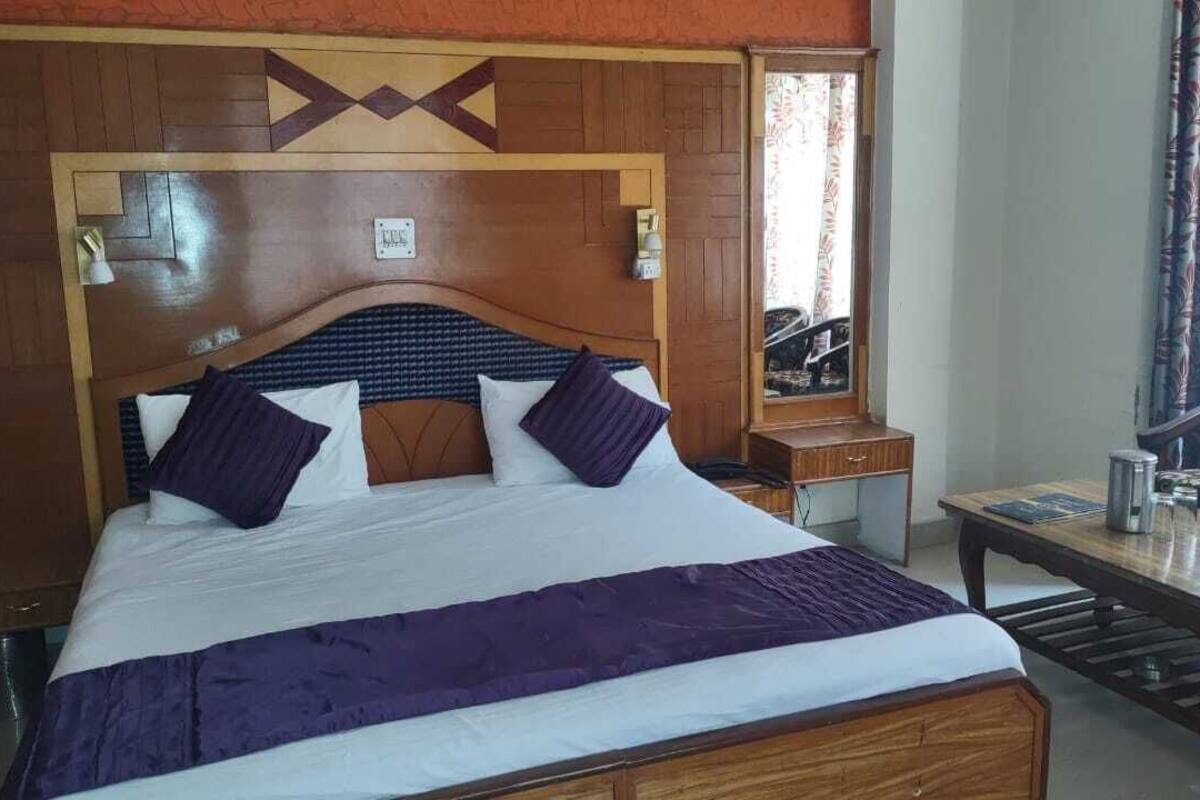 A hotel room with a wooden headboard, a bed with white and purple bedding, a small bedside table, and a wooden nightstand.