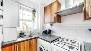 Een kleine keuken met houten kastjes, een wasmachine, een gasfornuis en een raam met een plant.