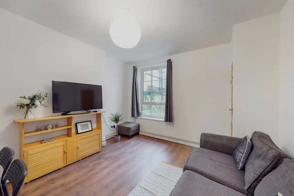 A living room with a flat-screen TV on a wooden console, a grey sofa, and a window with curtains.