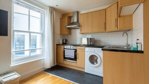 A compact kitchen with wooden cabinets, a built-in oven, a washing machine, and a window providing natural light. - Spaciously Renovated 2BD Flat - Camden (London)