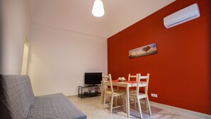 Una sala de estar con un sofá gris, una mesa de comedor con sillas, un televisor sobre un mueble bajo y un aire acondicionado en la pared.