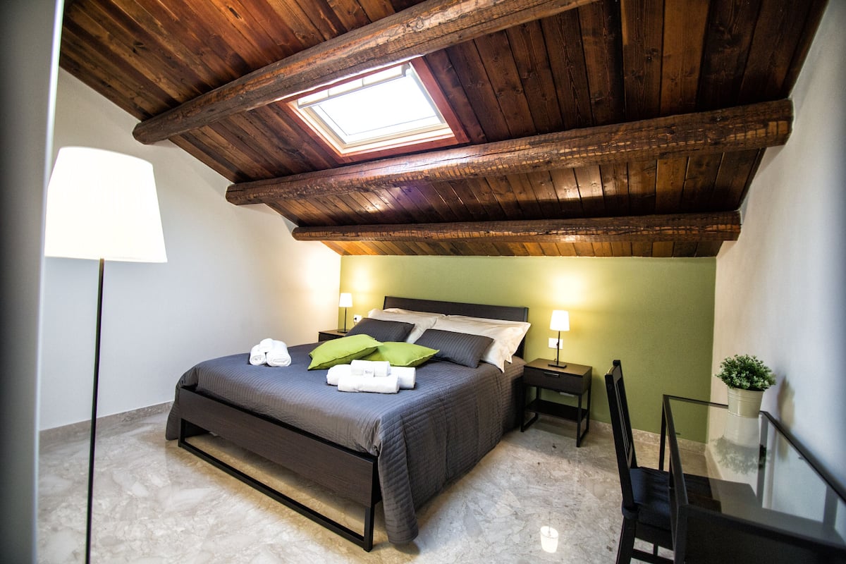 A bedroom with a skylight, a bed with pillows, a bedside table, a chair, and a small potted plant.
