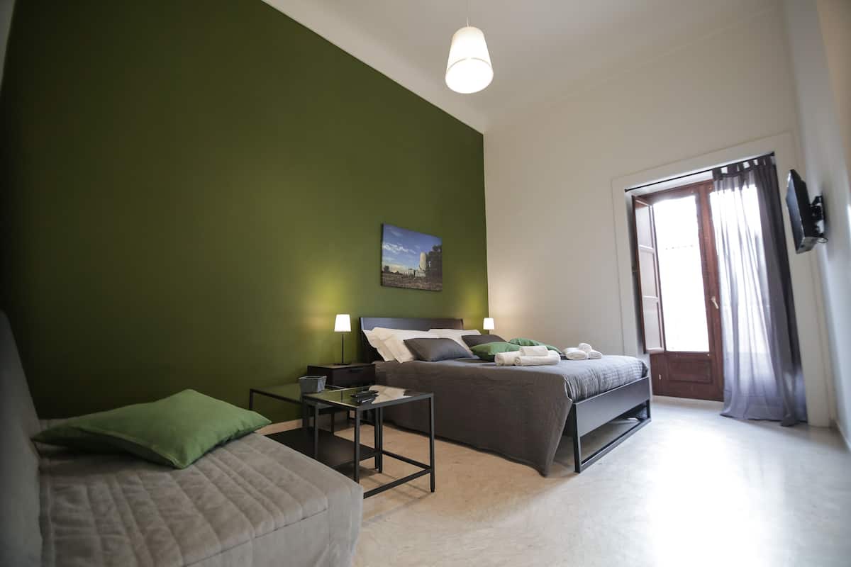 A bedroom with a bed, a green wall, a painting, a TV, and a window with curtains.