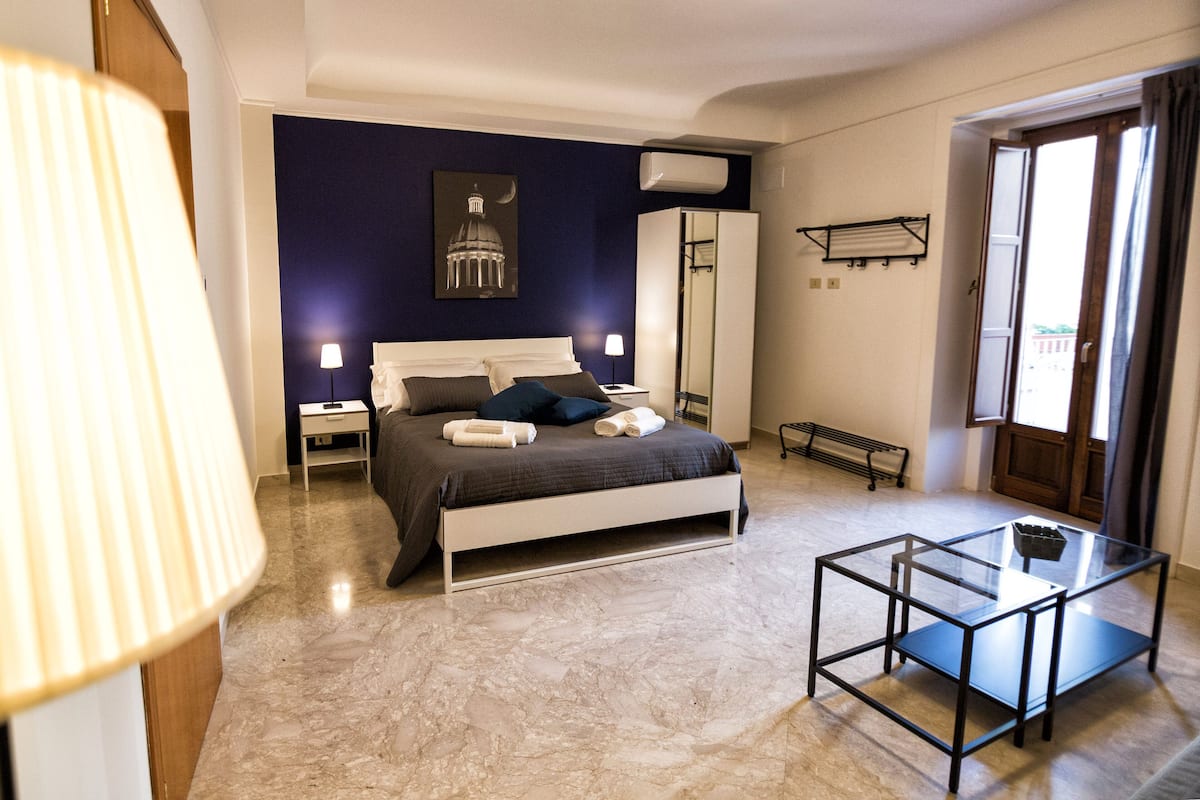 A bedroom with a bed, bedside tables, a glass table, a floor lamp, a ceiling fan, and a window.
