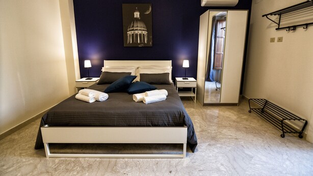 Una cama bien hecha con almohadas y toallas, una mesita de noche con lámpara, un aire acondicionado de pared y un espejo que refleja parte de la habitación.