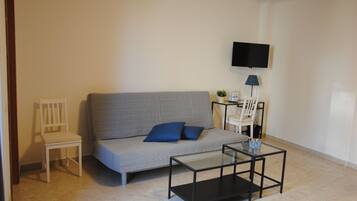 Un salon avec un canapé gris, un fauteuil blanc, une table basse en verre, un téléviseur fixé au mur et une petite table sur laquelle se trouvent une lampe et des bouteilles d’eau.