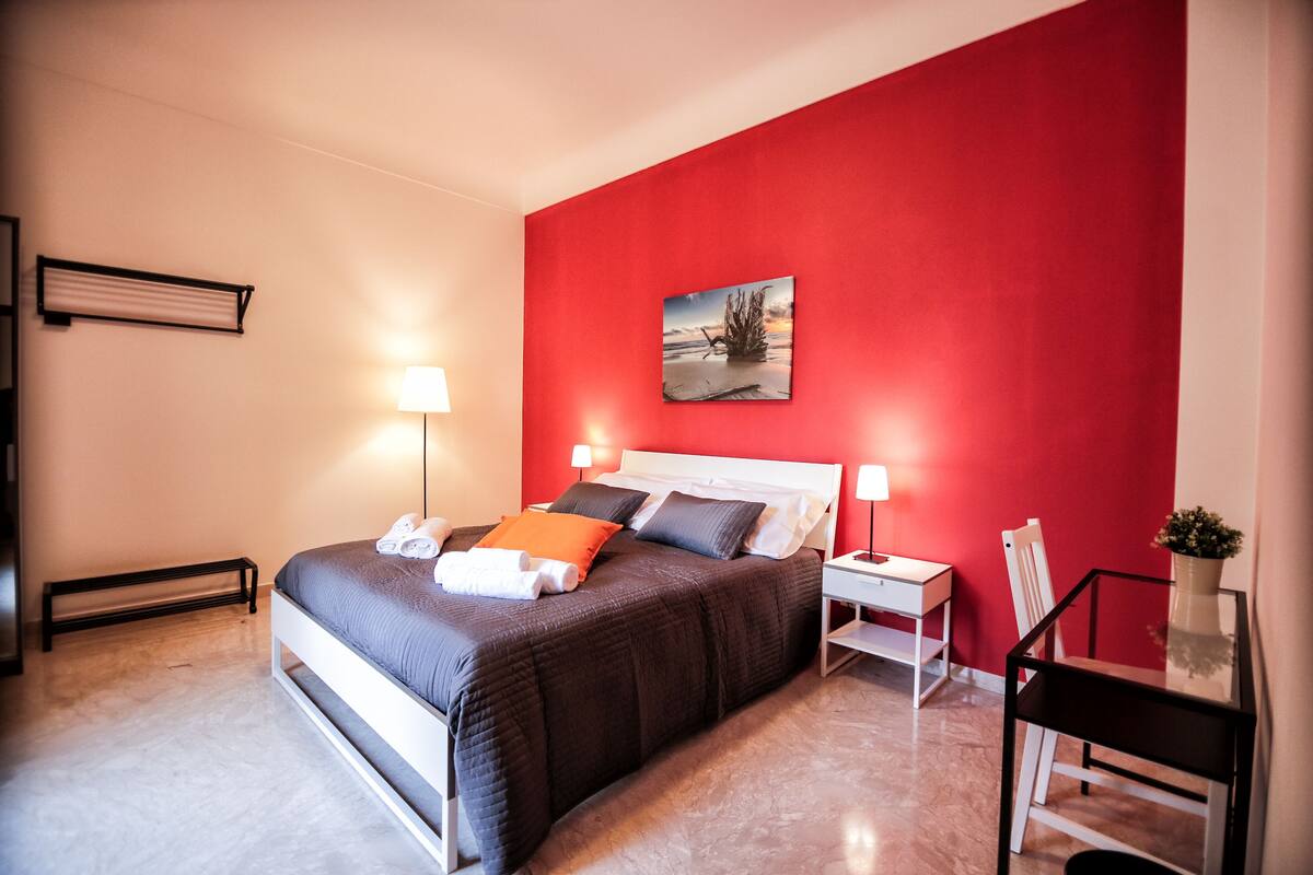 A modern bedroom with a red accent wall, a bed with a dark quilt, two bedside tables with lamps, a chair, and a small table with a plant.