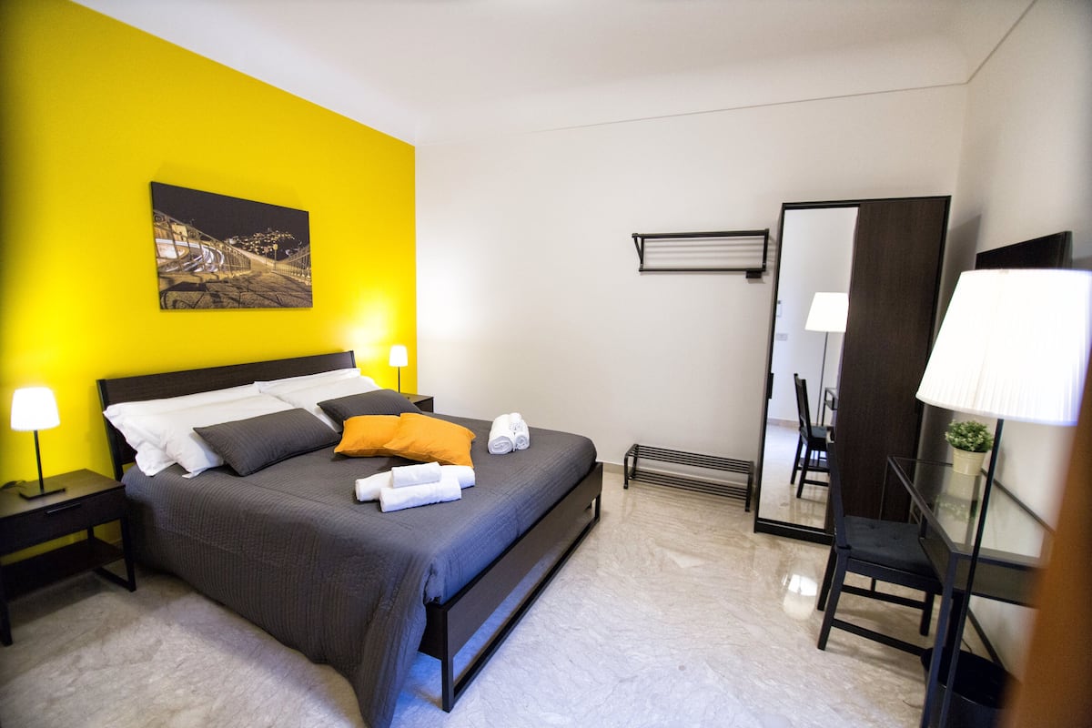 A modern hotel room with a large bed, a nightstand with a lamp, a mirror, and a chair.