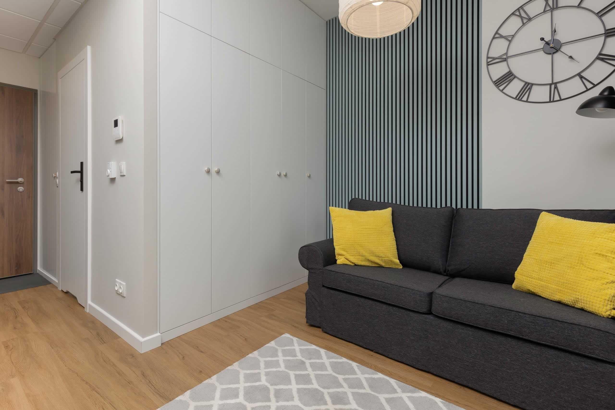 Een moderne woonkamer met een grijze bank, twee gele kussens, een kamerkleed met dessin, een wandklok en een verticaal gestreept wandpaneel.