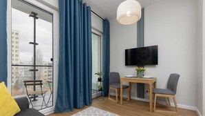 A modern living room with a sofa, a dining table with two chairs, a TV mounted on the wall, and a balcony with a view of buildings. - Compact Studio in Wola | Balcony | Air Conditioning (Warszawa)