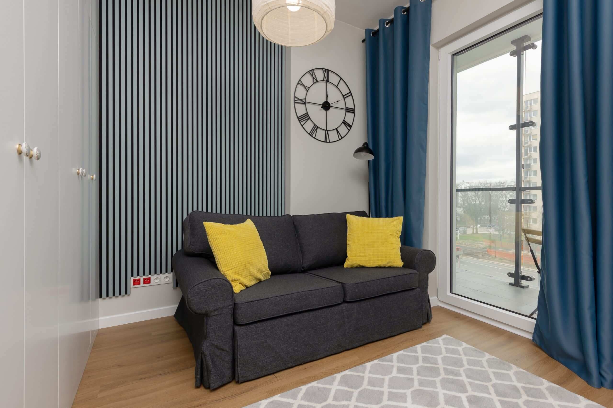 Un salon moderne avec un canapé gris, deux coussins jaunes, une horloge murale, un radiateur et une vue sur l’extérieur par une grande fenêtre.