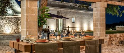 Un área de comedor al aire libre con un muro de piedra de fondo, vigas de madera y luces colgantes.