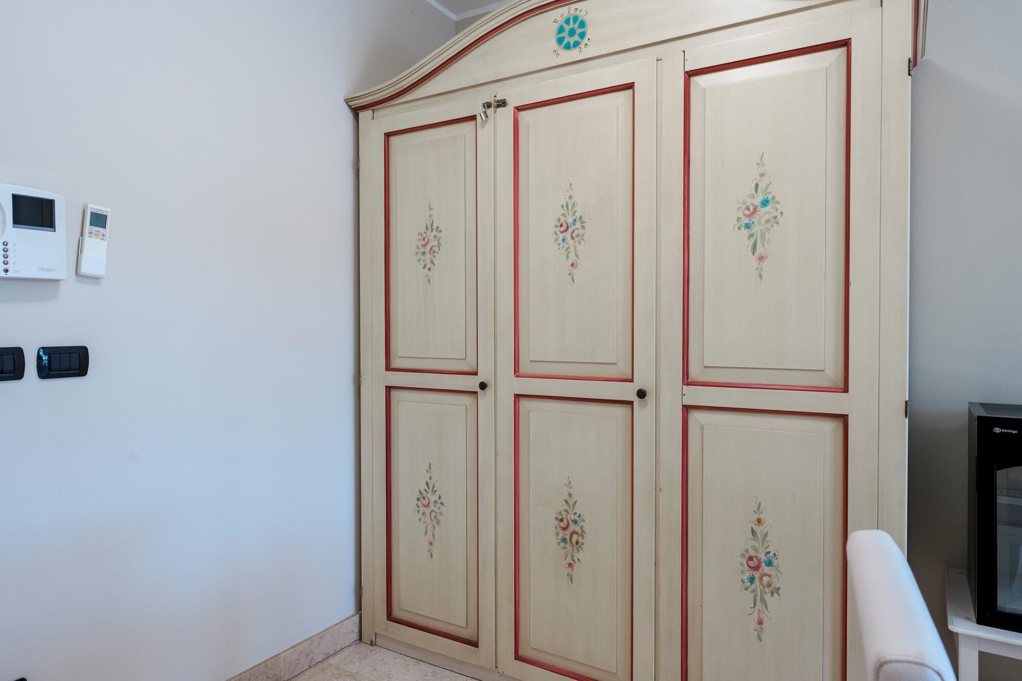 A room with a beige wardrobe featuring floral designs, a thermostat, and a television on a stand.