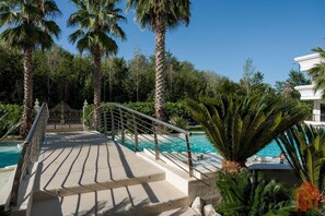 Un espace piscine avec un chemin en béton menant à la piscine, entouré de palmiers et d’un bâtiment blanc.