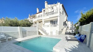 Una piscina con borde de baldosas blancas, una escalera que lleva a un balcón, y un edificio blanco con varios balcones y ventanas.