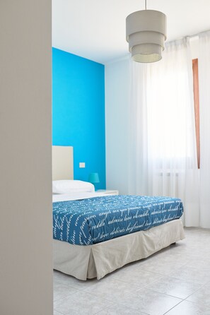 Een moderne hotelkamer met een blauwe accentmuur, een bed met een bedsprei met dessin, een nachtkastje met een lamp, en een groot raam met transparante gordijnen.