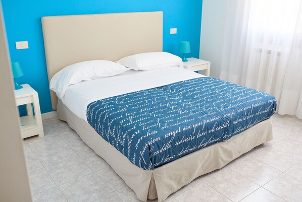 Een bed met een blauw-wit gebed en twee witte kussens, en een beige hoofdeinde in een kamer met blauwe muren en witte gordijnen.