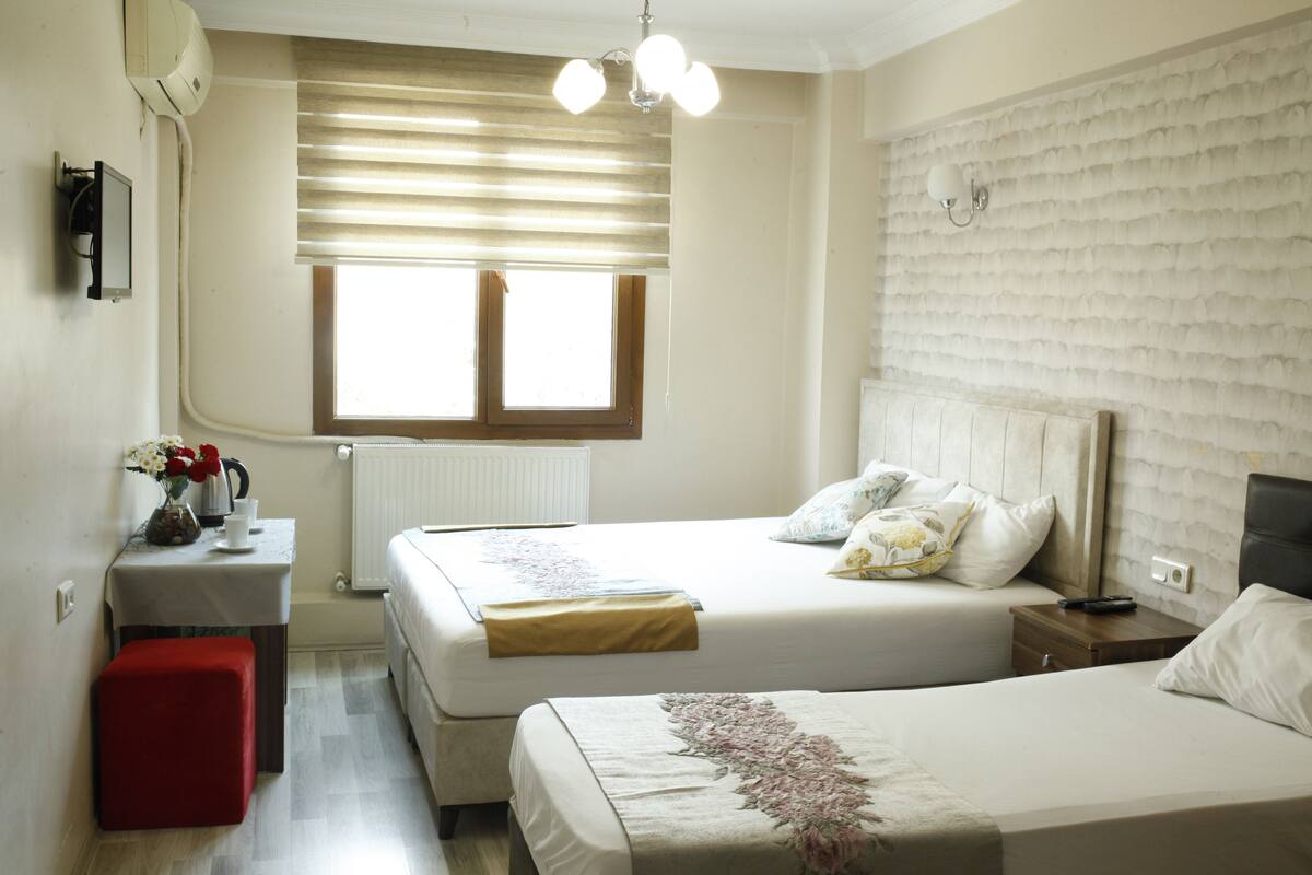 A bedroom with two beds, a small table with a vase of flowers, a red stool, a window with blinds, and a wall-mounted light fixture.