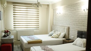 Habitación de hotel con dos camas, una mesita con un florero, un radiador y una ventana con persianas.