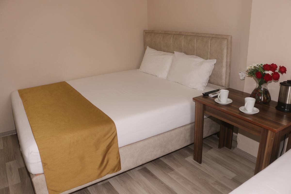 A neatly made bed with a white and mustard-colored bedspread, a wooden nightstand with a vase of red roses, and a coffee maker.