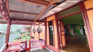 Eine überdachte Veranda mit einer Holzbank, einer Glasschiebetür und Blick auf einen Garten sowie ein Gebäude im Hintergrund.
