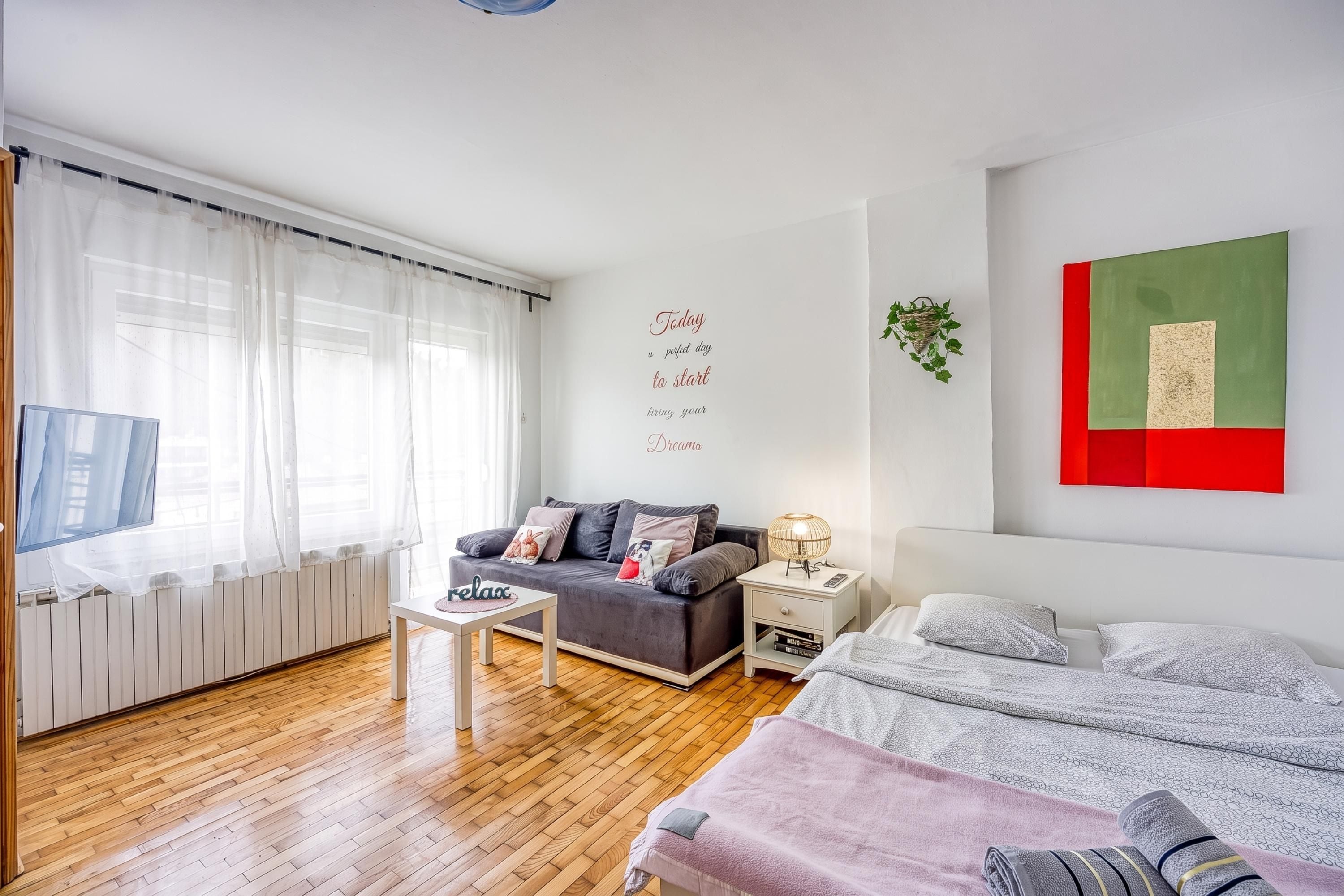A modern bedroom with a grey sofa, a bed with a grey and white patterned duvet, a small white bedside table with a lamp, and a wall with a green and red artwork.