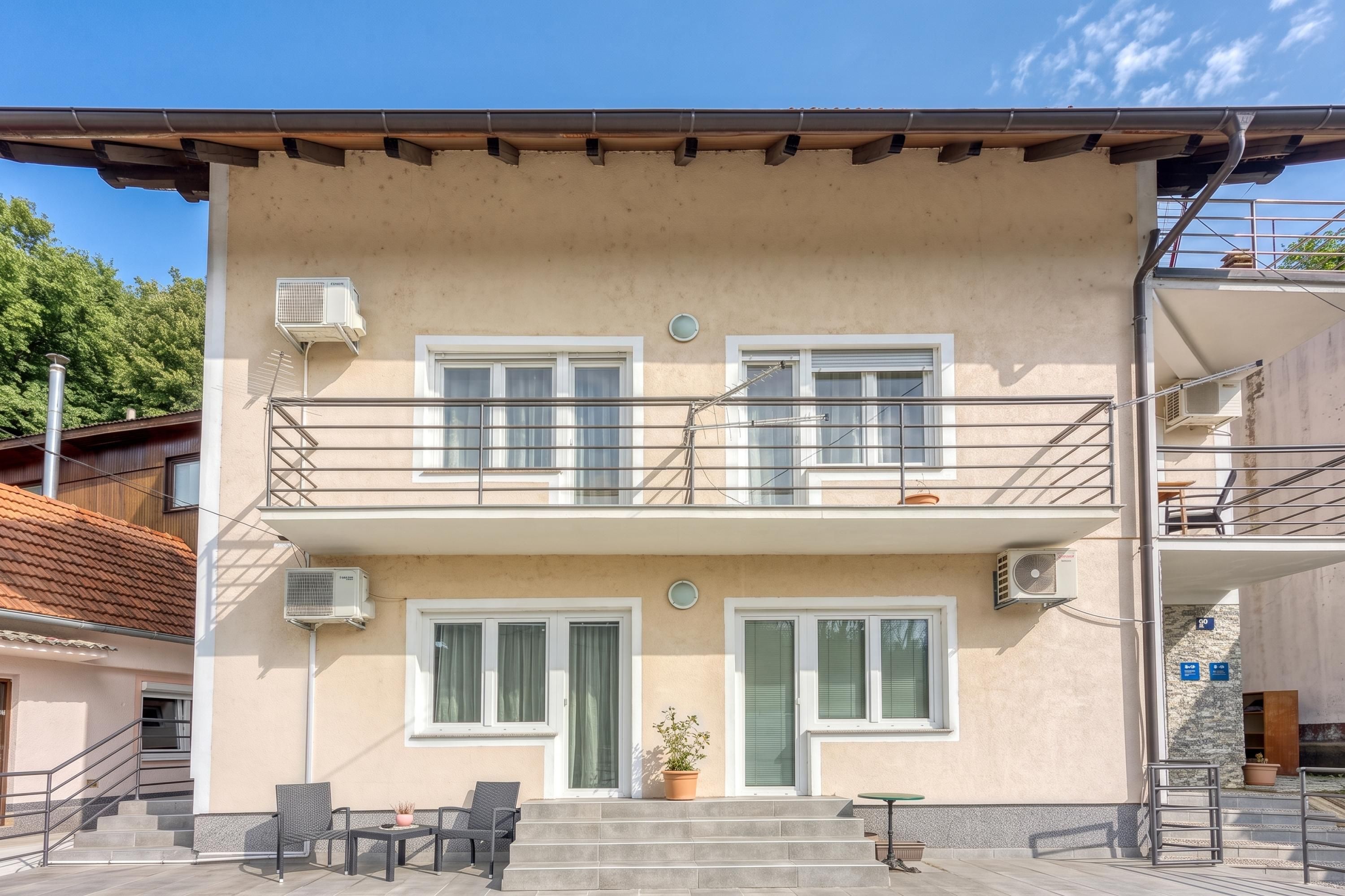 Un edificio de dos pisos con balcón, unidades de aire acondicionado y un pequeño espacio al aire libre con sillas y una mesa.