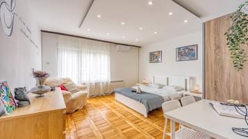 A modern bedroom with a wooden floor, a bed, a desk, and a chair. There is a window with curtains, a wall-mounted air conditioner, and two framed pictures.