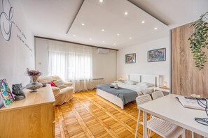 A modern bedroom with a wooden floor, a bed, a desk, and a chair. There is a window with curtains, a wall-mounted air conditioner, and two framed pictures. - Green Hill Zagreb Apartments (Zagreb)