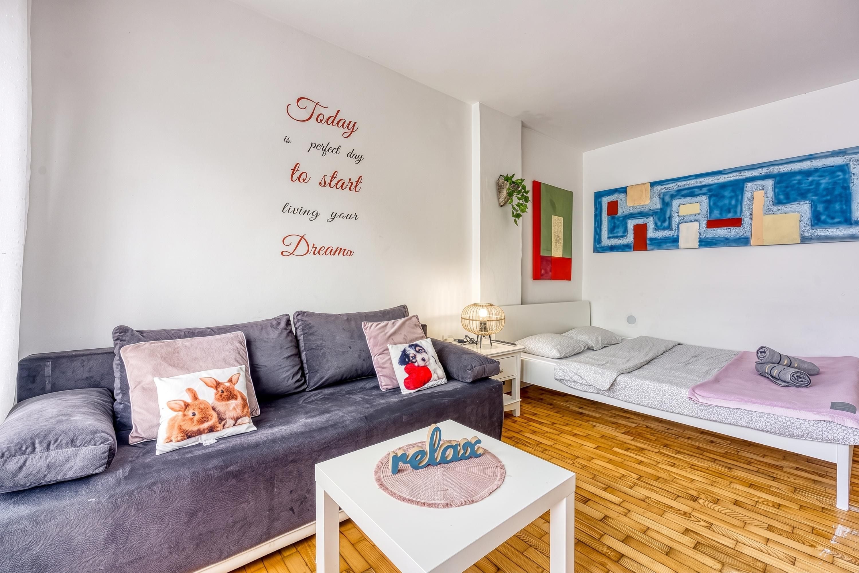 Uma sala de estar moderna com um sofá cinzento, uma mesa de centro branca e uma cama com manta cinza.