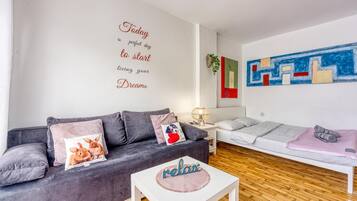 Uma sala de estar moderna com um sofá cinzento, uma mesa de centro branca e uma cama com manta cinza.
