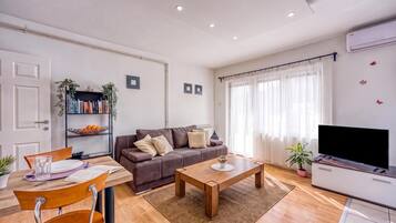 Una sala de estar moderna con un sofá, una mesa de centro y un televisor. Se observa una estantería con libros y objetos decorativos.