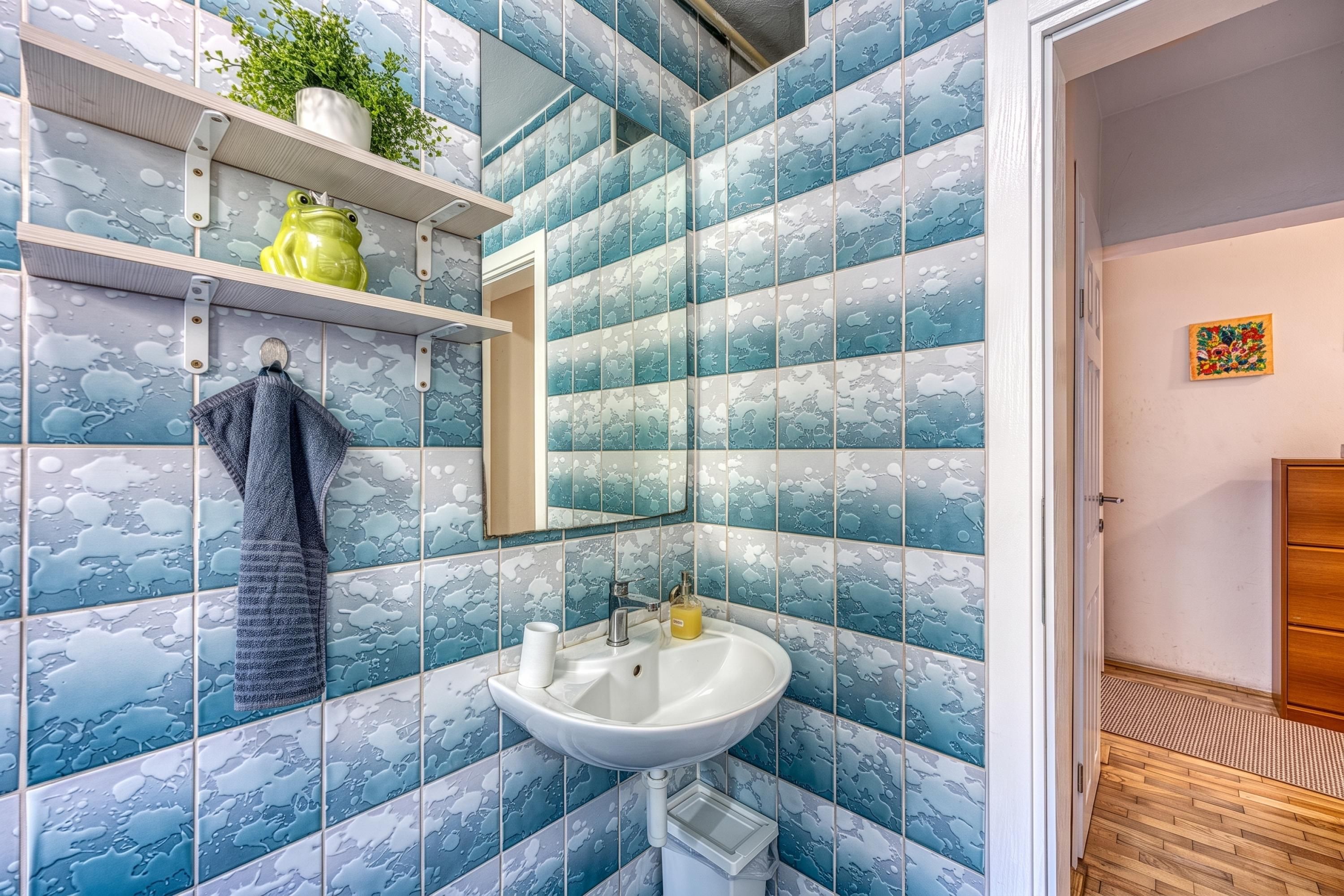 Um banheiro com azulejos estampados em azul, pia branca, toalha pendurada na parede e uma pequena prateleira com uma planta e um vaso amarelo.