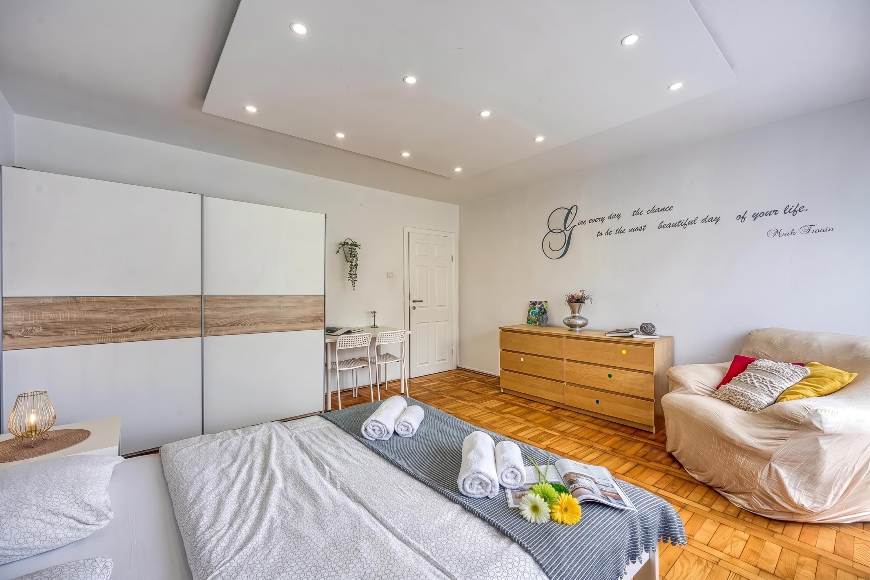 Ein modernes Schlafzimmer mit einem Bett, einem Sessel, einer Kommode und einer Wand mit motivierendem Text.