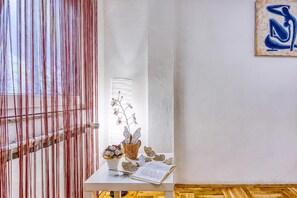 Una habitación con pared blanca, suelo de madera, ventana con persianas verticales, una pequeña mesa con un libro, un jarrón con flores y una obra de arte en la pared.