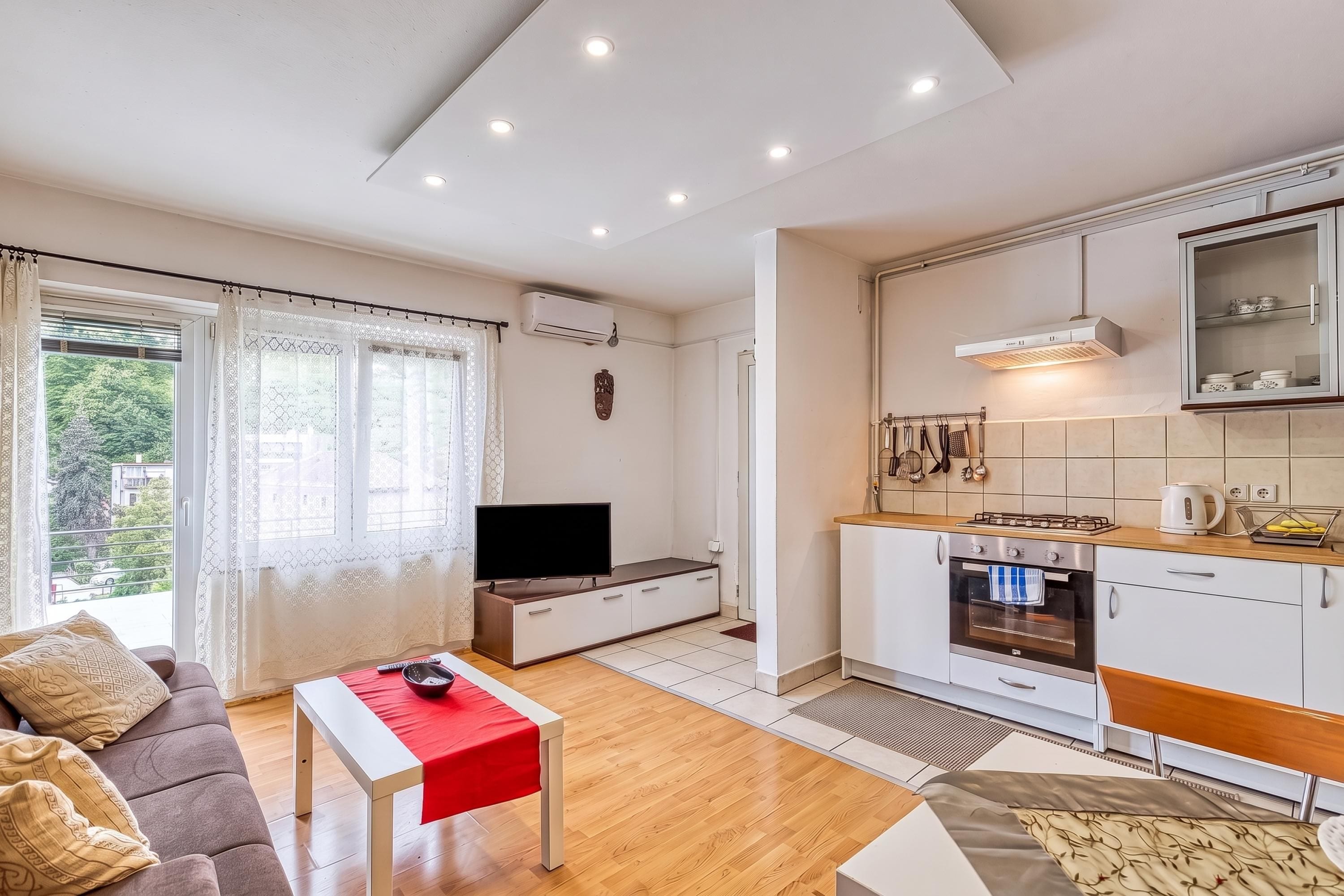 Uma sala de estar moderna com televisão de tela plana, sofá e mesa vermelha. A cozinha possui armários brancos, fogão e micro-ondas.