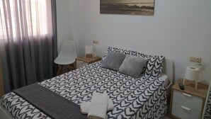 Ein Schlafzimmer mit Bett, Sessel, Nachttisch, Gemälde und Fenster mit Vorhängen.