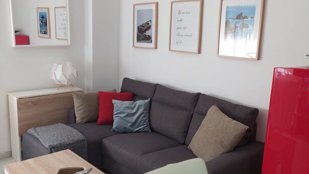 Ein modernes Wohnzimmer mit einem grauen Sofa, einem Holztisch und einem roten Kühlschrank. An den Wänden hängen gerahmte Bilder und ein kleiner weißer Tisch mit einem Korb.