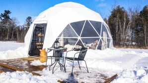 Lumipeitteinen iglu, jossa on läpinäkyvä kupoli, puiset ovet ja pieni pöytä tuoleineen.